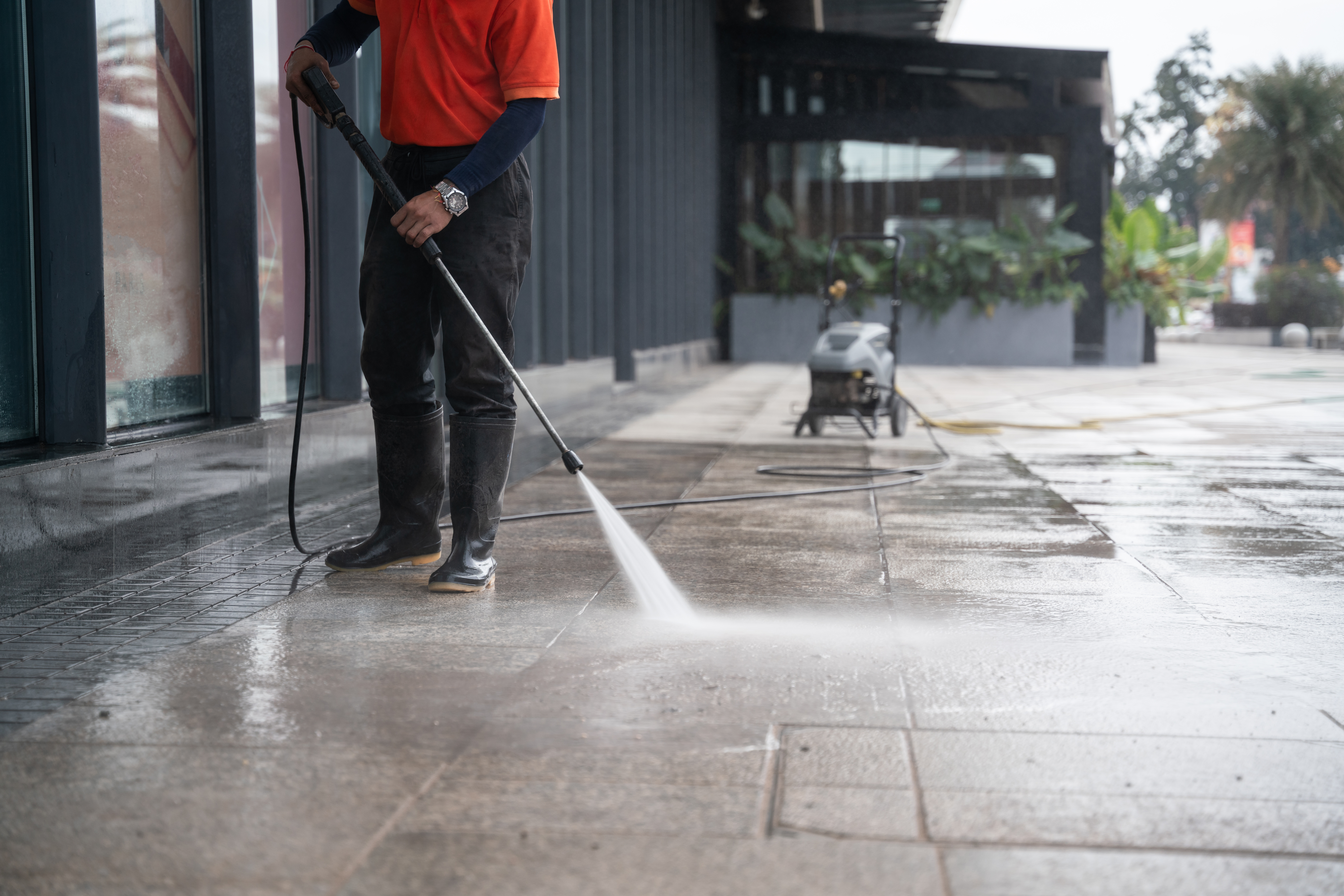man in cleaning clothes and rubber boots pressure washing pavement outside office building for commercial spring cleaning.