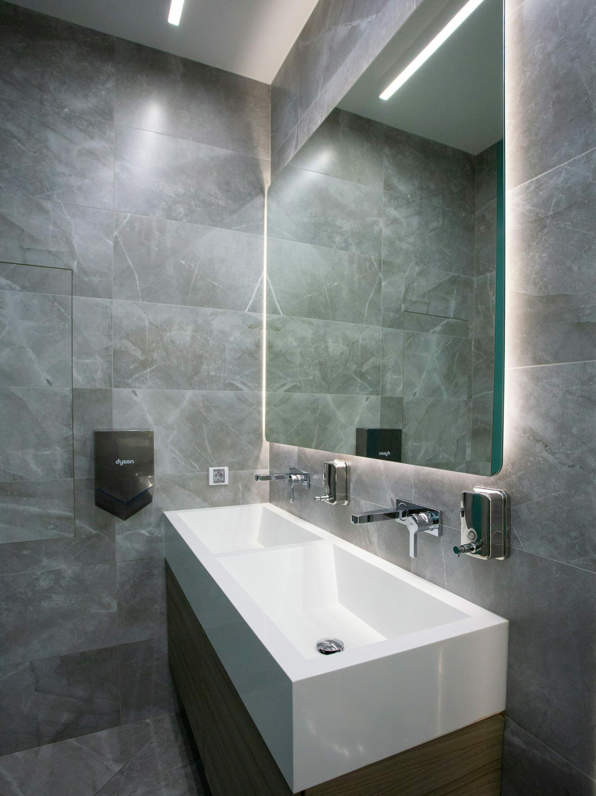 a modern public bathroom with grey tile, a mirror with backlighting, and double white sunken-in sinks with chrome facets/fixtures.